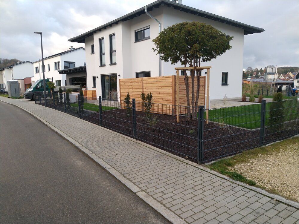 Modernes zweistöckiges Haus mit schwarzem Drahtzaun und hölzernem Sichtschutzzaun im Vorgarten. Gepflasterter Gehweg und Blick auf die Straße.