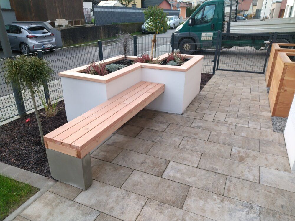 Eckbank mit integrierten Pflanzkästen, weiße Wand, Holzbank und -verkleidung, gepflasterte Terrasse, grüner LKW im Hintergrund.