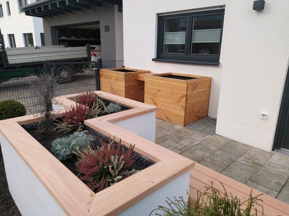 Pflanzkästen und hölzerne Pflanzgefäße in der Nähe eines Hauses mit Gartengestaltung; im Hintergrund ist ein Lastwagen zu sehen.