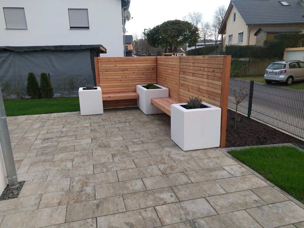 Eine Holzbank und Pflanzkästen auf einer Steinterrasse, im Hintergrund ein hölzerner Sichtschutzzaun, ein Auto und Häuser.