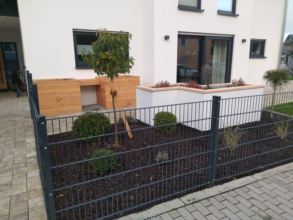 Modernes Hausäußeres mit angelegtem Vorgarten, schwarzem Zaun und verschiedenen Sträuchern und kleinen Bäumen.