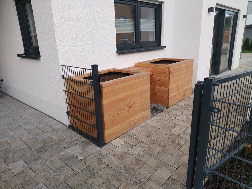 Zwei hölzerne Pflanzkästen an einem weißen Haus mit gepflasterter Terrasse, in der Nähe eines Metallzauns.