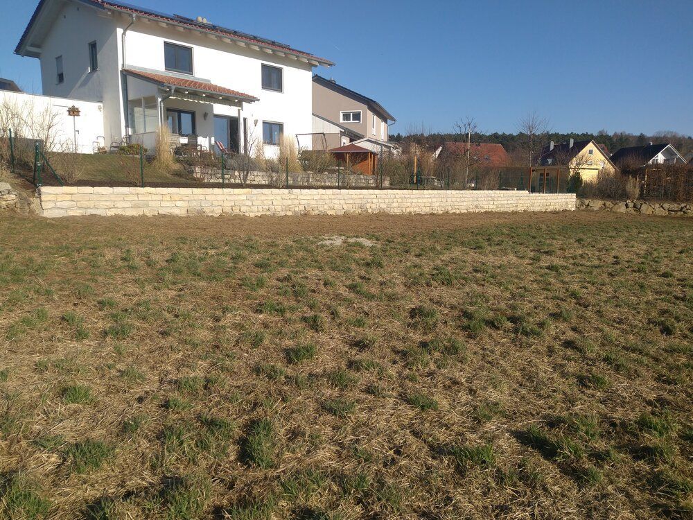 Rasenbewachsener Hof vor einem weißen Haus mit einer steinernen Stützmauer und kleinen Bäumen im Hintergrund.