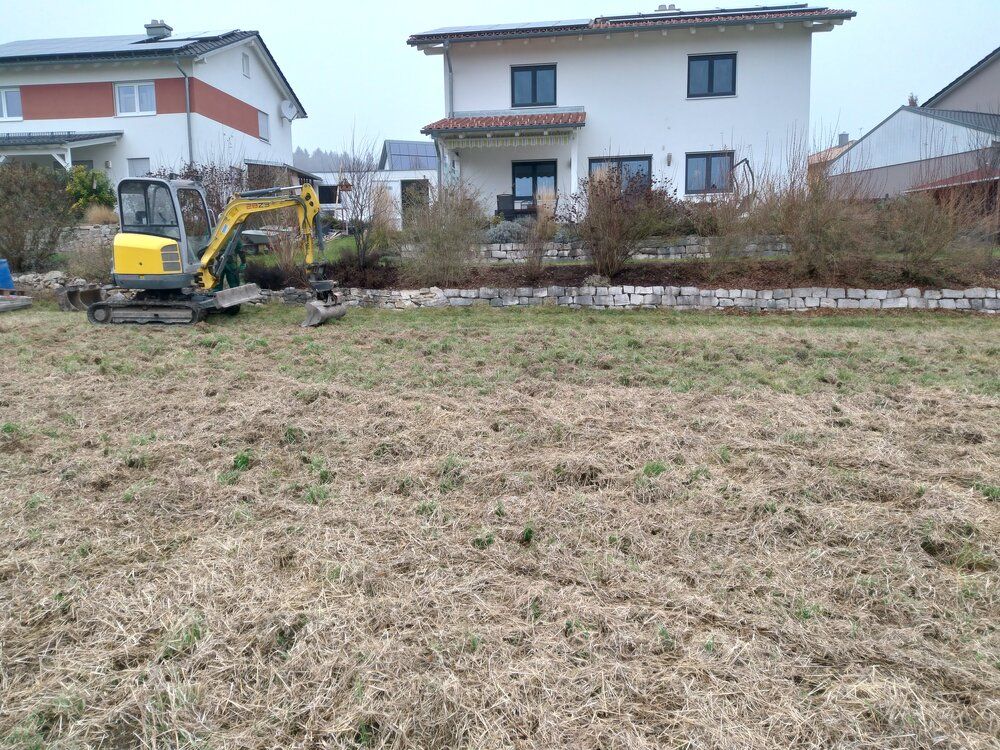 Ein gelber Bagger in einem grasbewachsenen Hof, in der Nähe einer Backsteinmauer und eines zweistöckigen weißen Hauses mit Sträuchern.