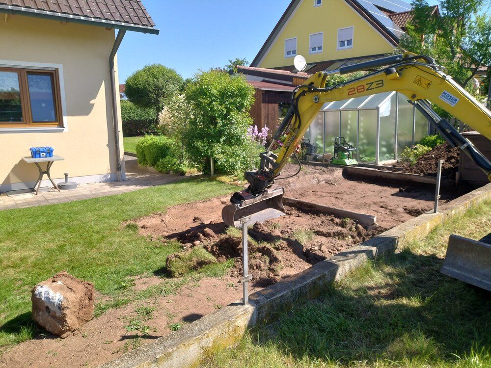 Ein Bagger gräbt in einem Hinterhof und hebt Erde ab. Grünes Gras umgibt eine Betonbegrenzung und das Haus.