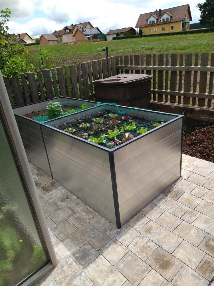 Zwei erhöhte Metallbeete mit Pflanzen auf einer Terrasse, im Hintergrund ein Holzzaun, Häuser an einem Hang.