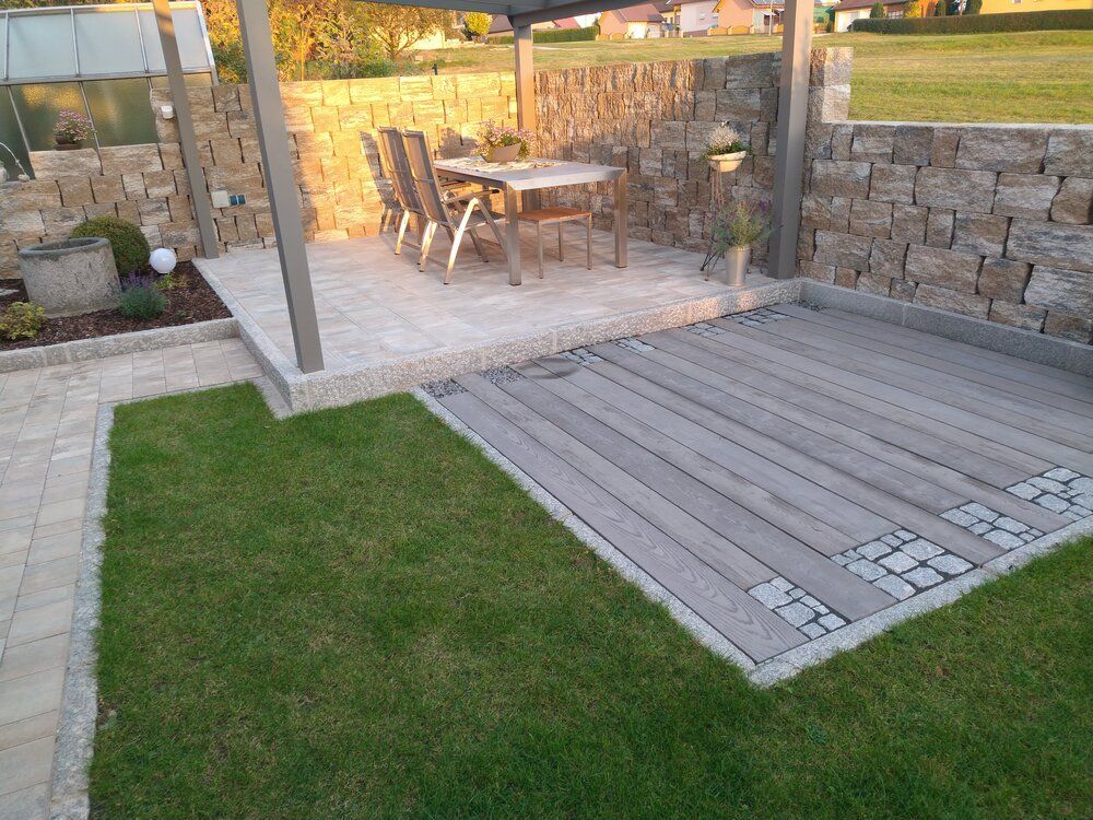 Terrasse mit Holzdeck und Steinmauern, Tisch und Stühle unter einer Pergola, grüner Rasen.