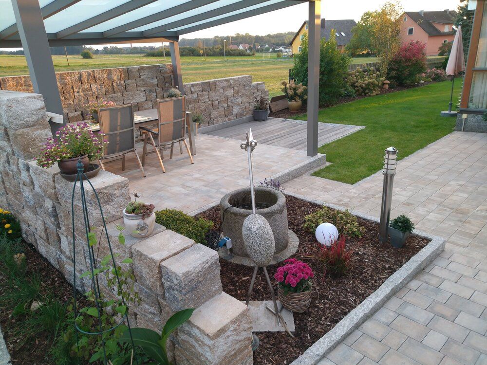 Steinterrasse mit Sitzgelegenheiten, Gartenbeeten und Wasserspiel unter einem überdachten Bereich, eingebettet in einen Garten.