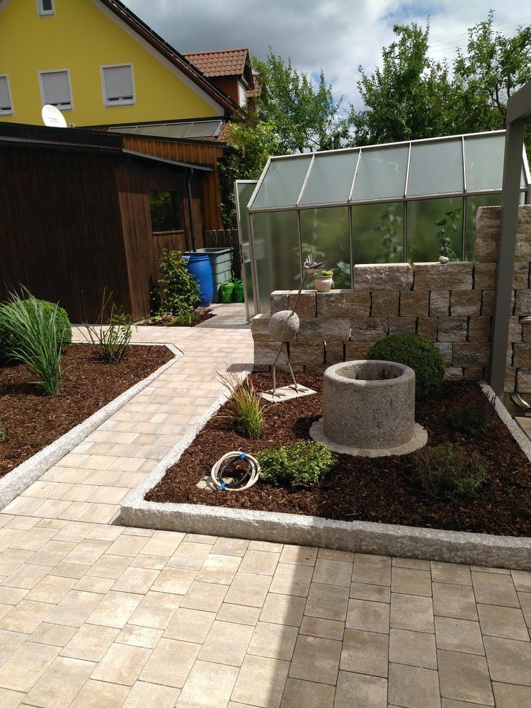 Ein Pfad führt zu einem kleinen Garten mit Gewächshaus, Steinbrunnen und Mulch.