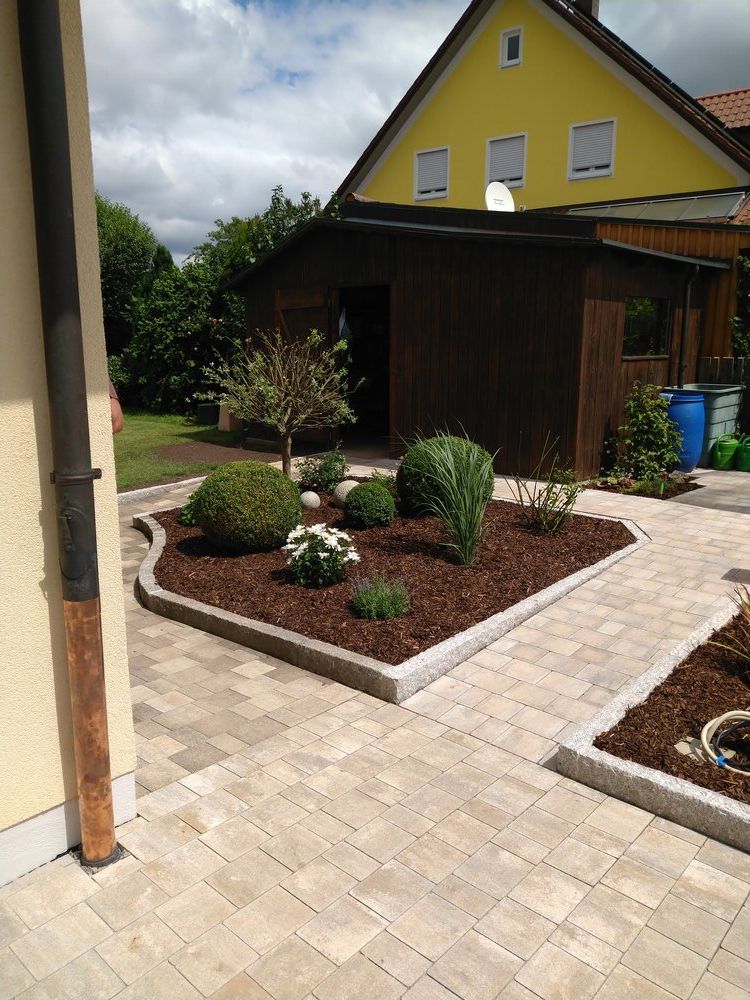 Eine mit Ziegeln gepflasterte Terrasse mit einem angelegten Beet. Kleine Sträucher und Blumen in einem mit Mulch bedeckten Beet. Im Hintergrund ein gelbes Haus.