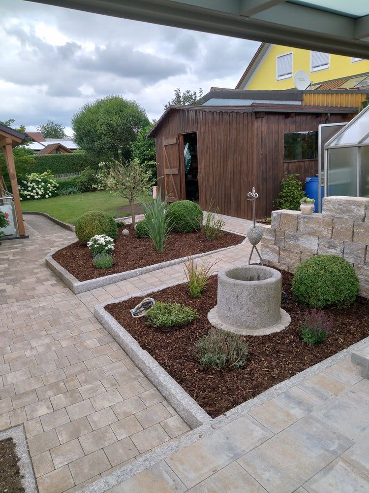 Ein gepflegter Hinterhof mit einer Ziegelterrasse, mit Mulch bedeckten Beeten, einem Springbrunnen und einem Holzschuppen.