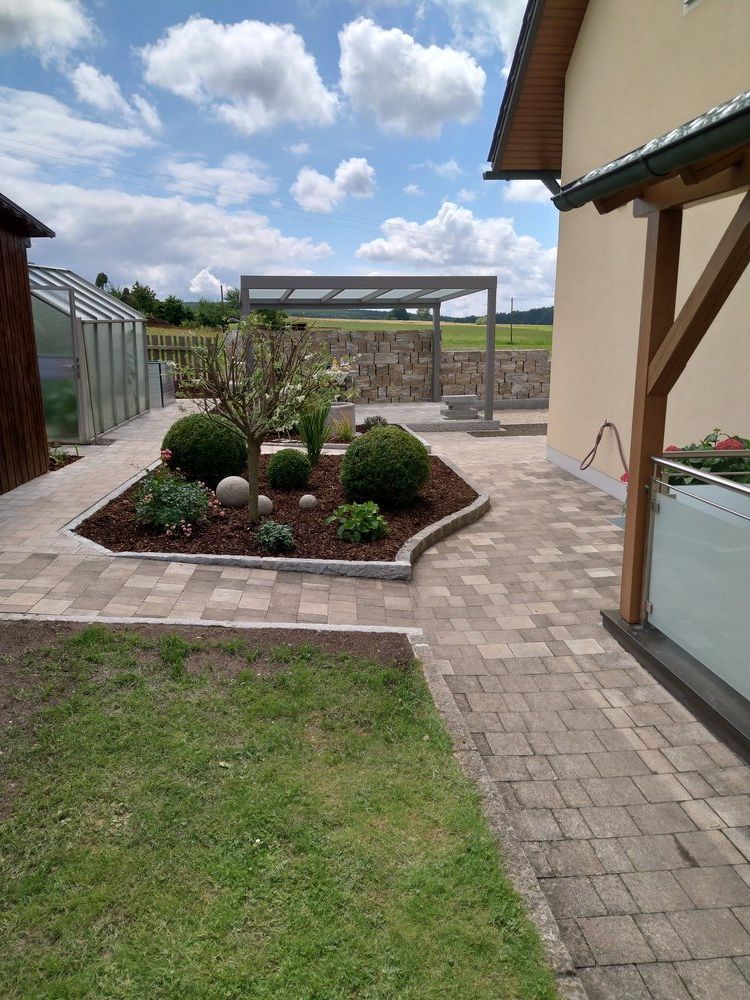 Ein schön angelegter Garten mit Steinterrasse, üppigem Grün und einer Pergola unter einem teilweise bewölkten Himmel.