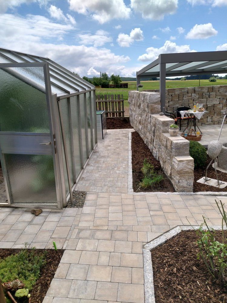 Ein Weg aus hellen Pflastersteinen führt vorbei an einem Gewächshaus und einer Steinmauer mit einem schattigen Terrassenbereich vor einer grünen Landschaft unter bewölktem Himmel.