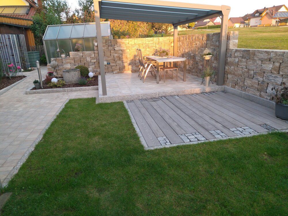 Terrasse im Freien mit Steinmauern, Holzdeck, Rasen und Pergola mit Tisch und Stühlen.