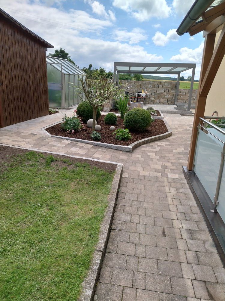 Eine mit Ziegeln gepflasterte Terrasse mit Blumenbeet, Gewächshaus und Holzkonstruktion unter einem teilweise bewölkten Himmel.