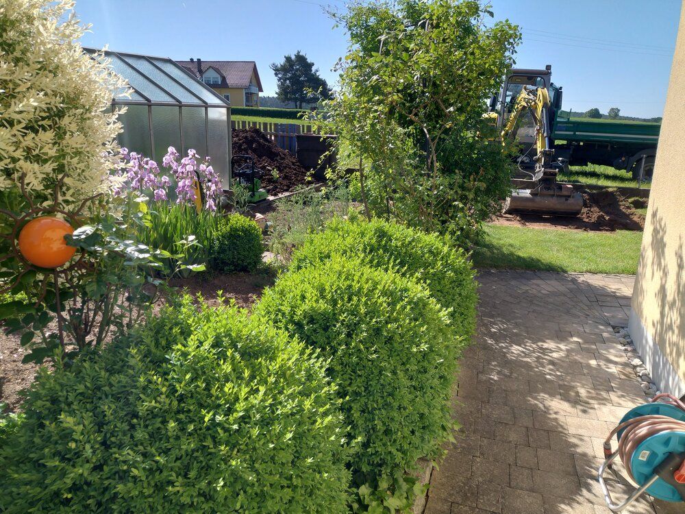 Grüne Hecken säumen einen Backsteinweg neben einem Garten mit blühenden Pflanzen und einem kleinen Gewächshaus; im Hintergrund ist ein Bagger zu sehen.