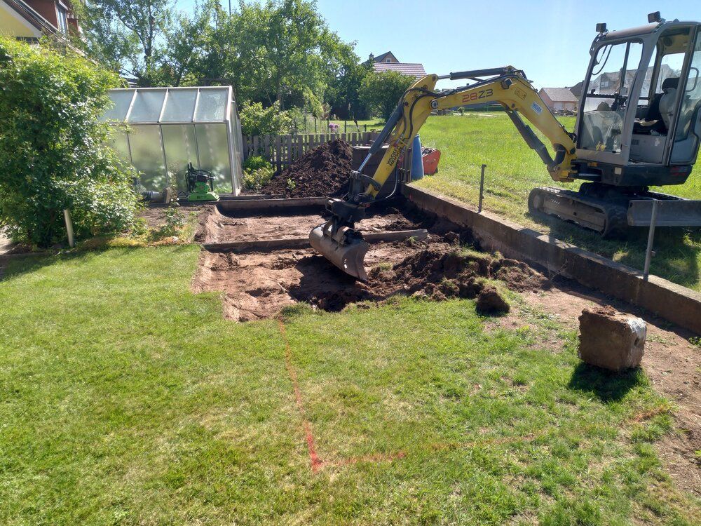 Ein Minibagger gräbt an einem sonnigen Tag in einem Hinterhof, in der Nähe eines Gartens und eines Gewächshauses.