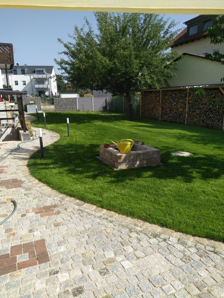 Hinterhof mit Kopfsteinpflasterweg, grünem Rasen, Feuerstelle, gestapeltem Brennholz und Bäumen unter blauem Himmel.