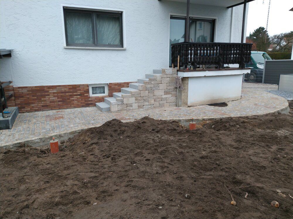 Steinstufen führen zu einer Veranda mit dunklem Geländer. Ein Kiesweg schlängelt sich um einen unbefestigten Weg.