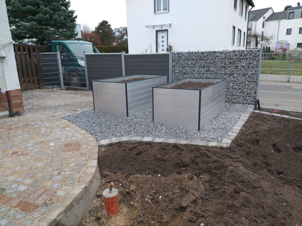 Zwei erhöhte Metall-Gartenbeete auf Kies, neben einem Stein- und grauen Zaun vor einem Haus.