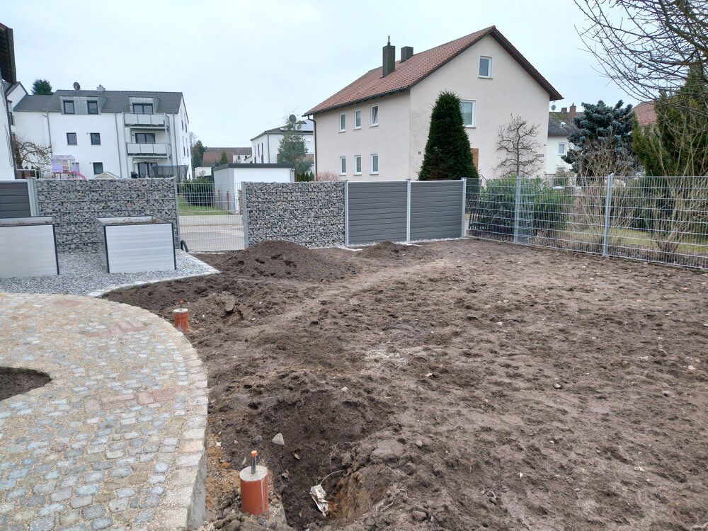 Baustelle mit aufgeschütteter Erde, neuem Zaun und Gabionenwänden aus Stein in der Nähe von Häusern an einem bewölkten Tag.