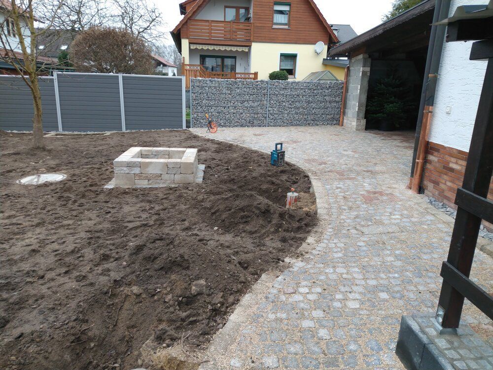 Ein Hinterhof mit Feuerstelle, Erde, einem Steinweg, einem grauen Zaun und einer Garage.