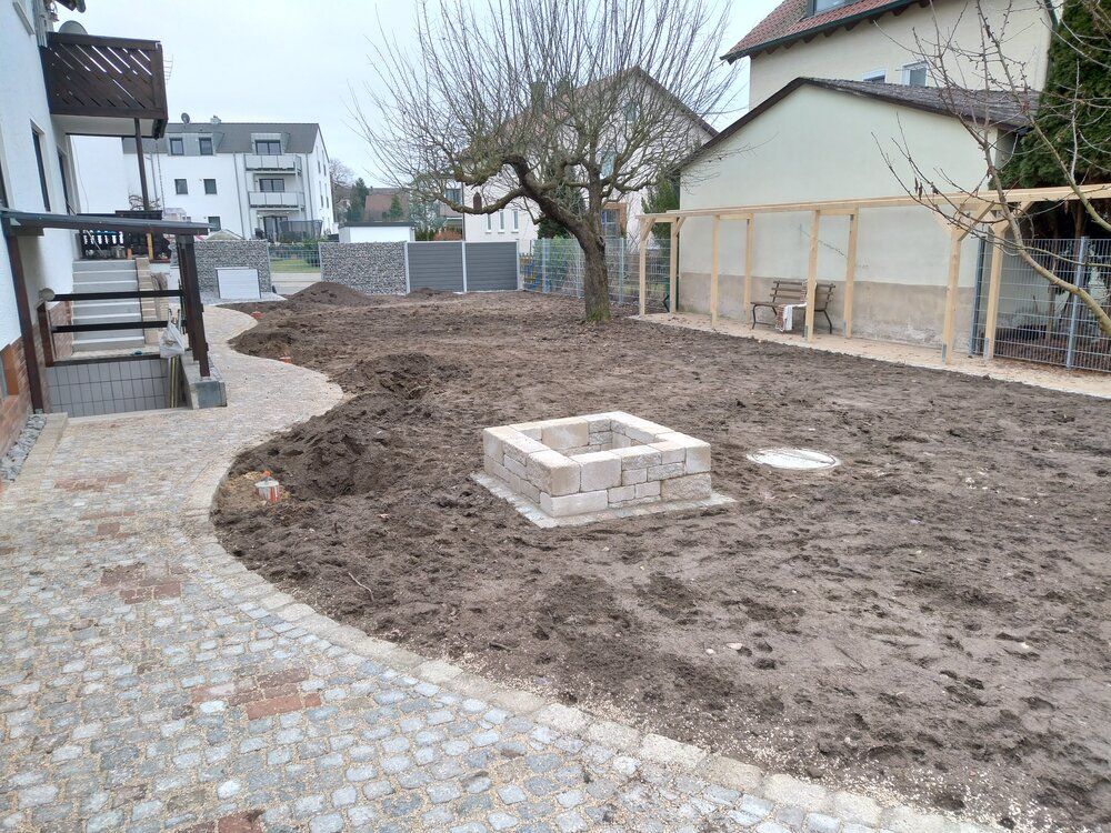Schlammiger Hinterhof mit steinerner Feuerstelle, Pfad und einem unfertigen Gebäude. Im Hintergrund kahle Bäume und Häuser.