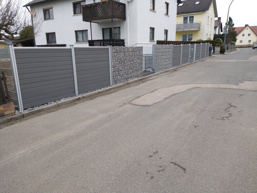 Grauer Zaun entlang einer Straße, mit horizontalen Paneelen in Holzoptik und einem Steinabschnitt. Weißes Haus im Hintergrund.