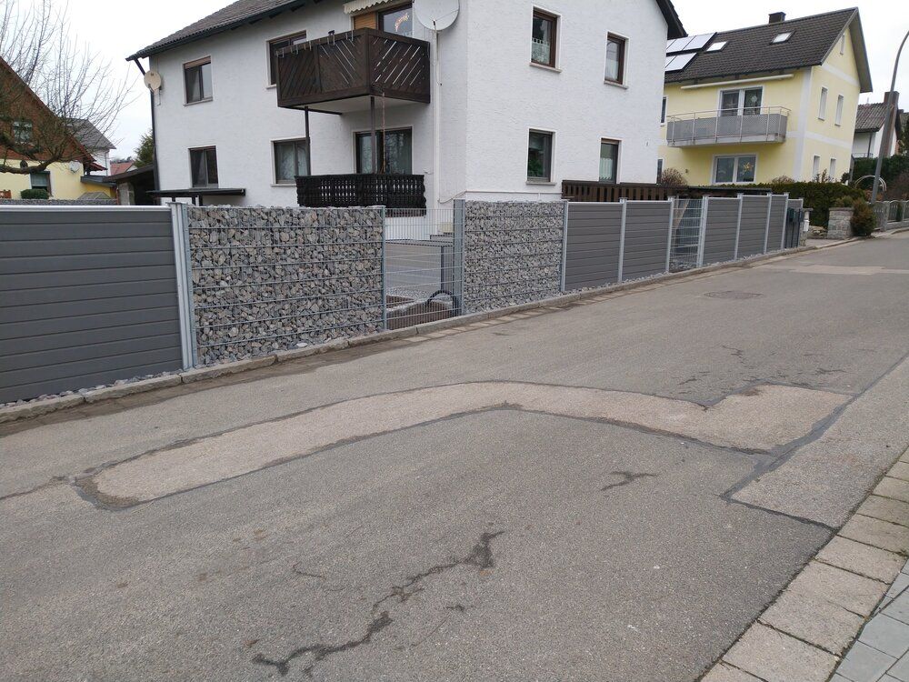 Graue Steinmauern säumen eine Straße, im Hintergrund sind Häuser zu sehen.