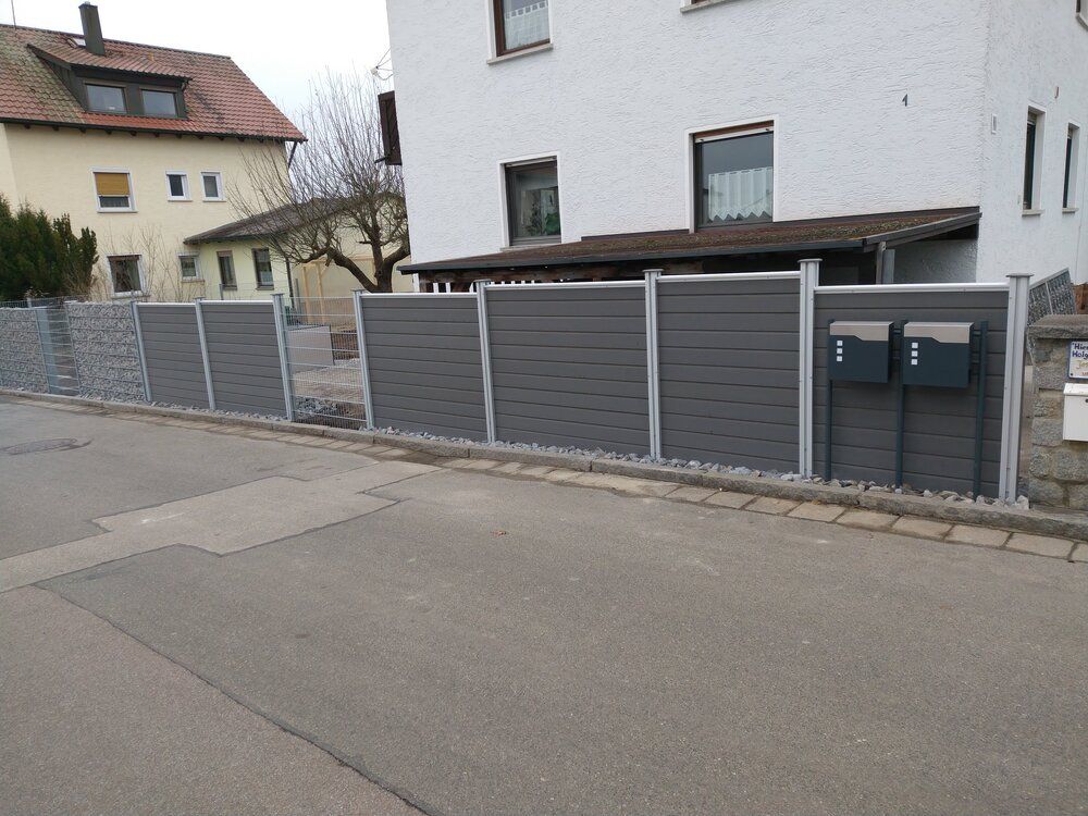 Grauer Verbundzaun mit Steinelementen und Briefkästen vor einem Haus an einer Straße.