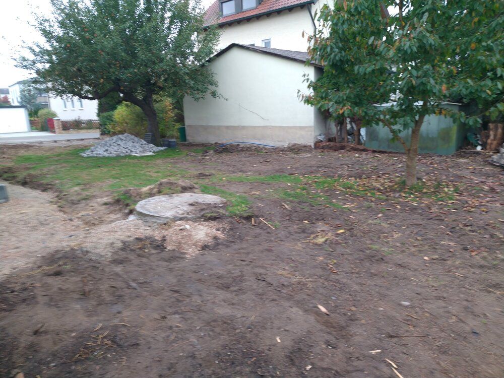 Hinterhof mit aufgewühltem Boden, einem Versorgungsschachtdeckel und einem Haus im Hintergrund.
