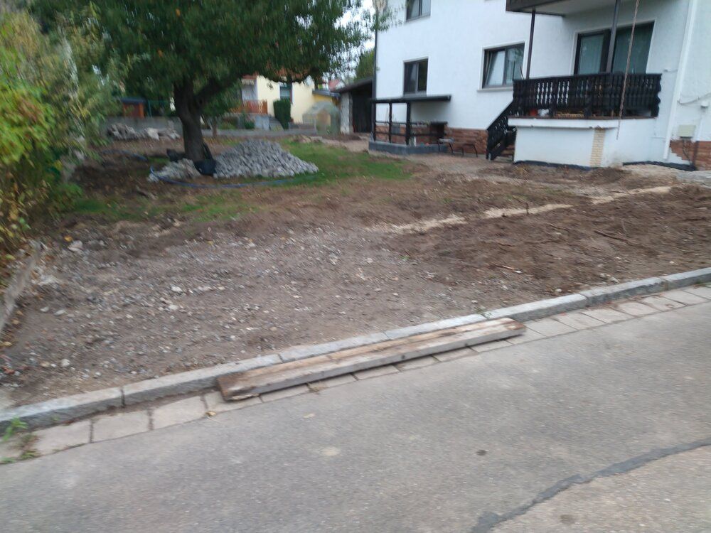 Ein mit Kies und unbewachsenem Erdreich bedeckter Hof grenzt an eine asphaltierte Straße und ein weißes zweistöckiges Gebäude.