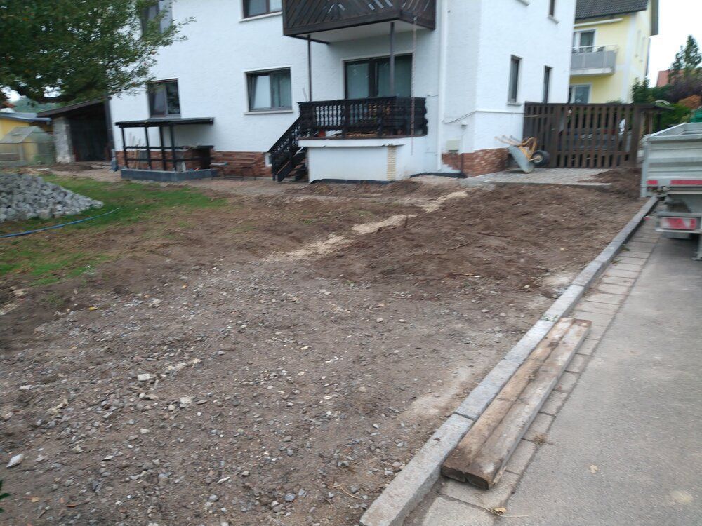 Ein Garten, der neben einem weißen Haus mit Balkon und grauem Gehweg für die Landschaftsgestaltung vorbereitet wird.