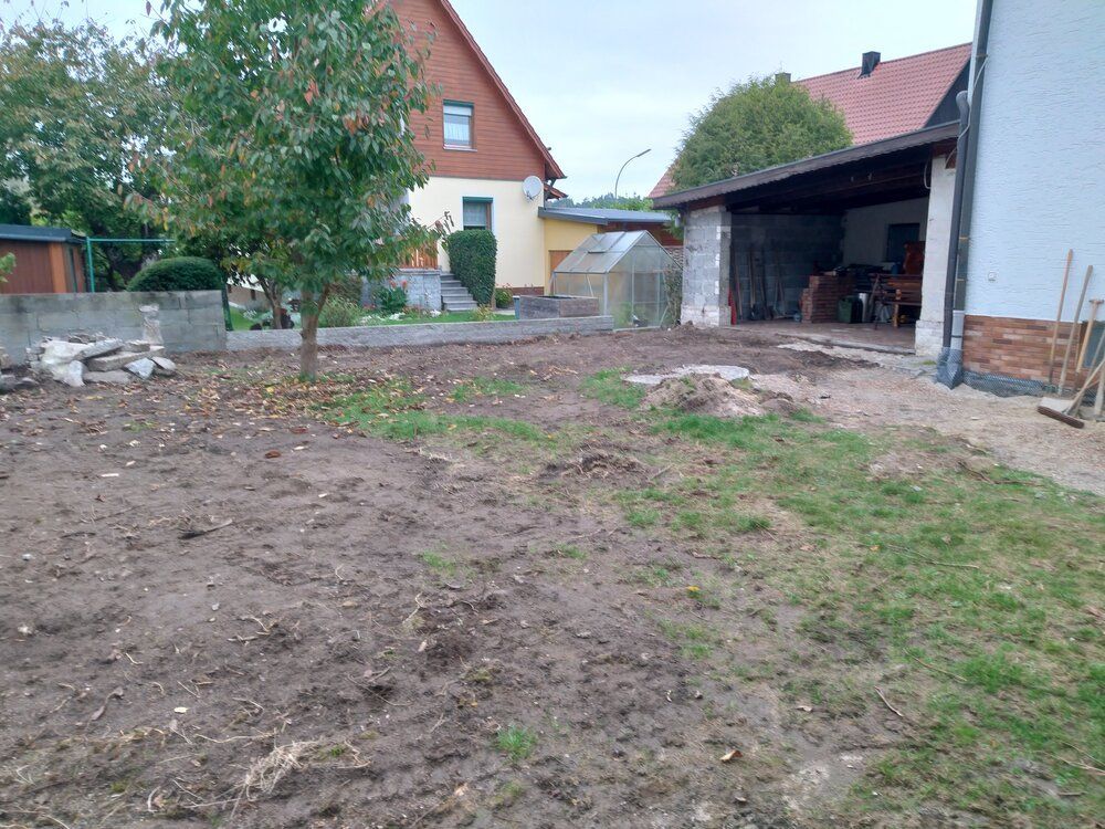 Schlammiger Hof mit spärlichem Gras, Schuppen, Baum und Gebäuden im Hintergrund. Bewölkter Tag.
