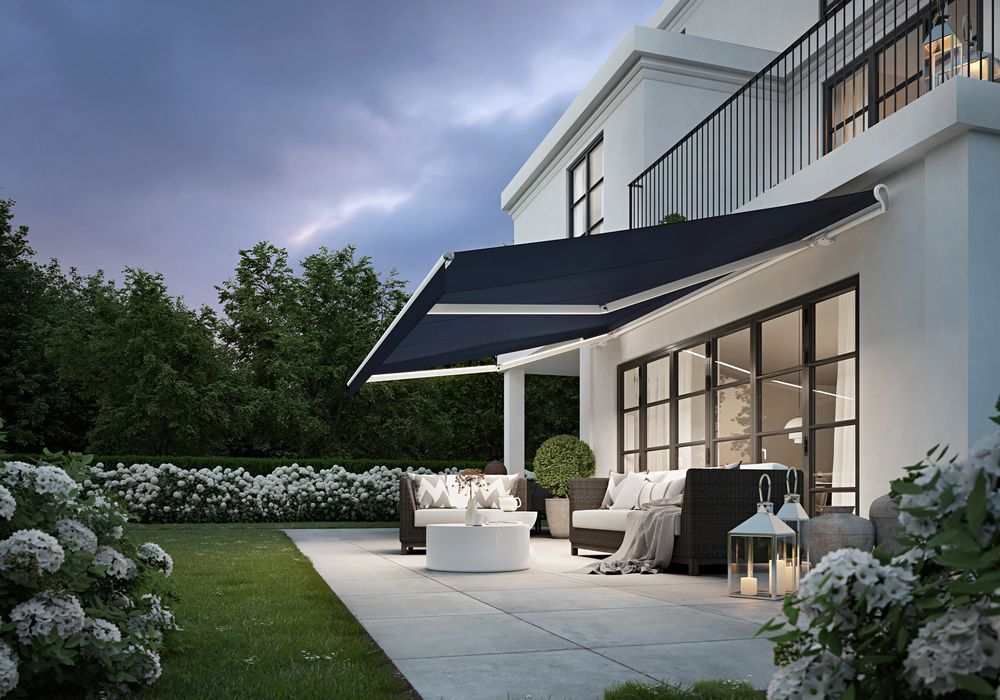 Weißes Haus mit Terrassenmöbeln unter einer Markise in der Abenddämmerung. Grüner Rasen und weiße Blumen im Vordergrund.