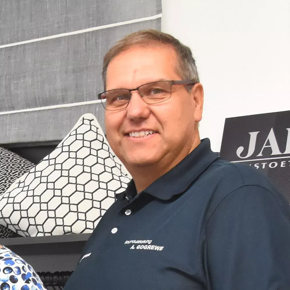 Ein Mann mit Brille und dunkelblauem Poloshirt lächelt und posiert neben Zierkissen.