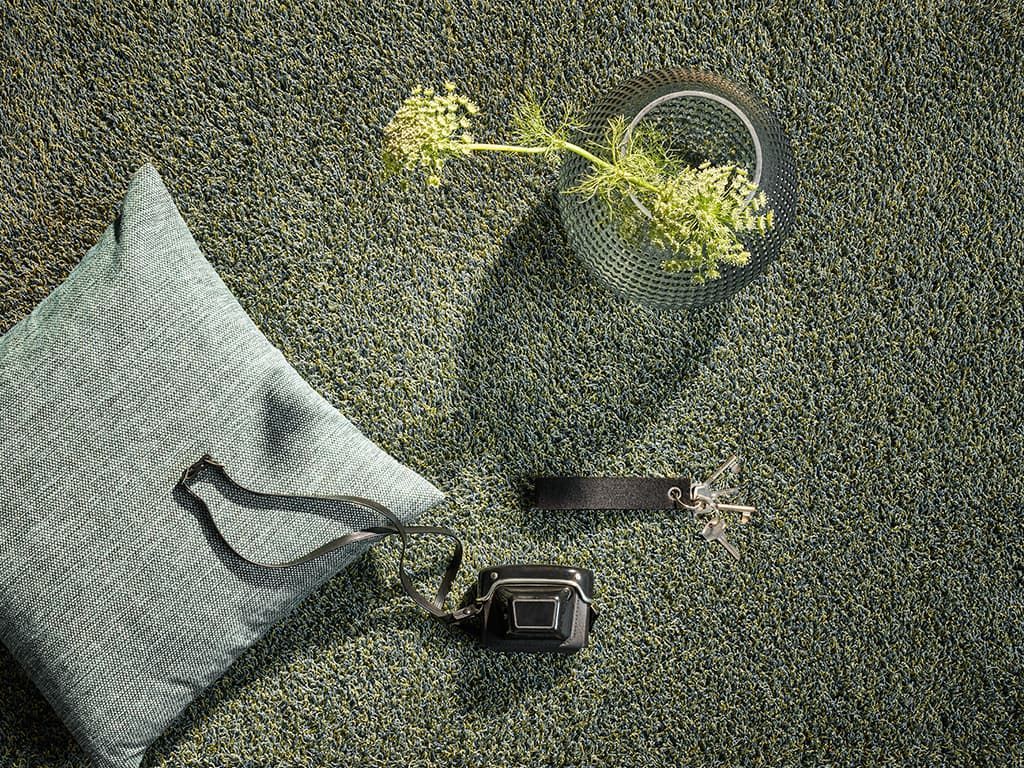 Kissen, Blumenvase, Gürtel und kleine schwarze Tasche auf einem strukturierten grünen Teppich.