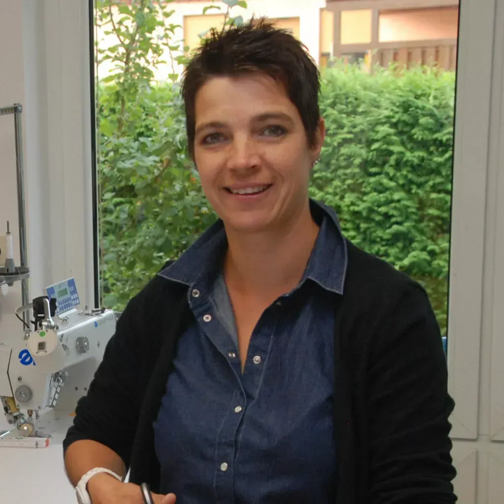 Frau in blauem Hemd und schwarzem Pullover lächelt an einer Nähmaschine, im Hintergrund ein Fenster.