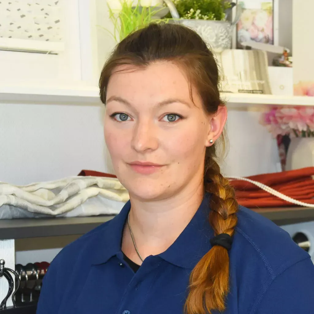 Eine Frau mit geflochtenem Haar und blauem Hemd lächelt leicht; sie steht drinnen in der Nähe von Regalen mit Dekorationsartikeln.