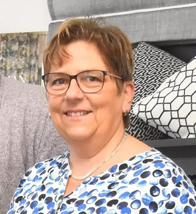 Frau mit Brille lächelt, trägt ein blau-weiß gemustertes Hemd und befindet sich in einer Wohndekorationsumgebung.