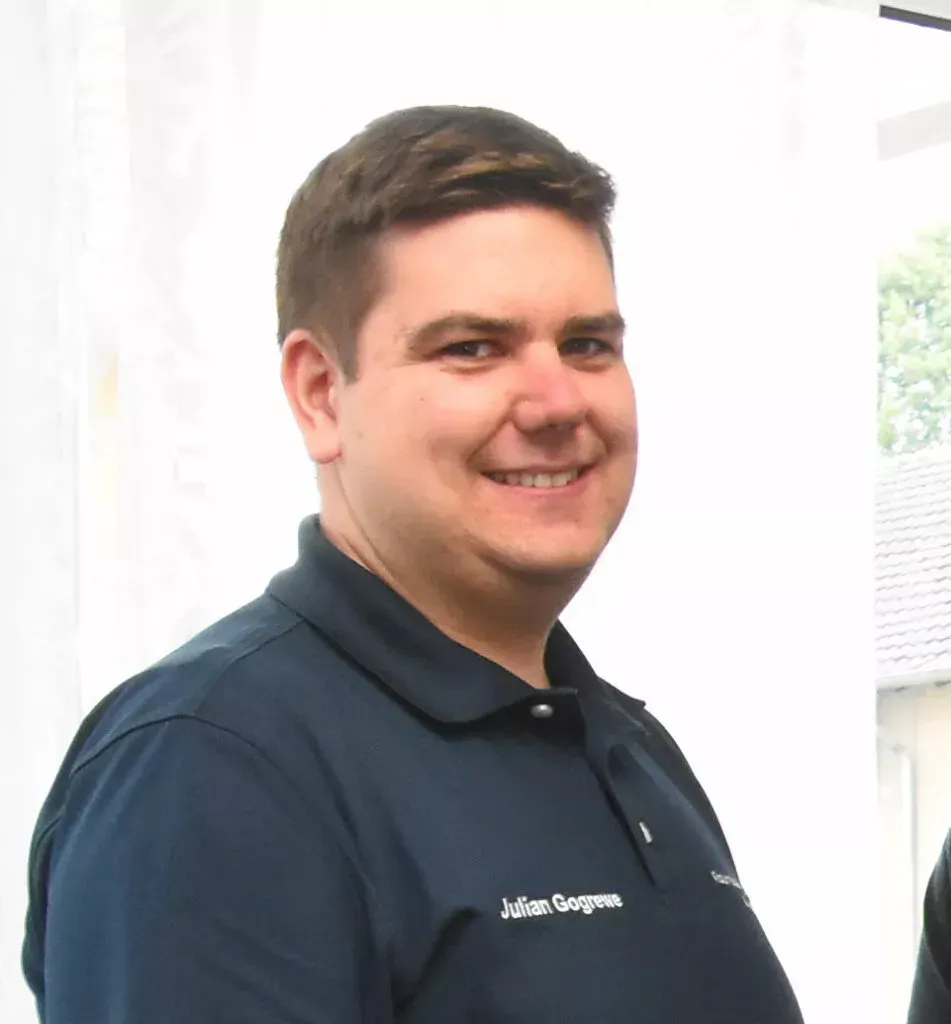 Ein lächelnder Mann in einem dunkelblauen Poloshirt in einem Zimmer mit Fenster vor weißem Hintergrund.