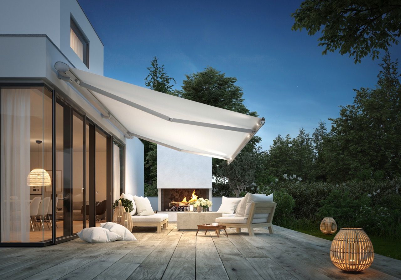 Terrasse in der Abenddämmerung mit ausfahrbarer Markise, weißen Möbeln, Kamin und Laternen.