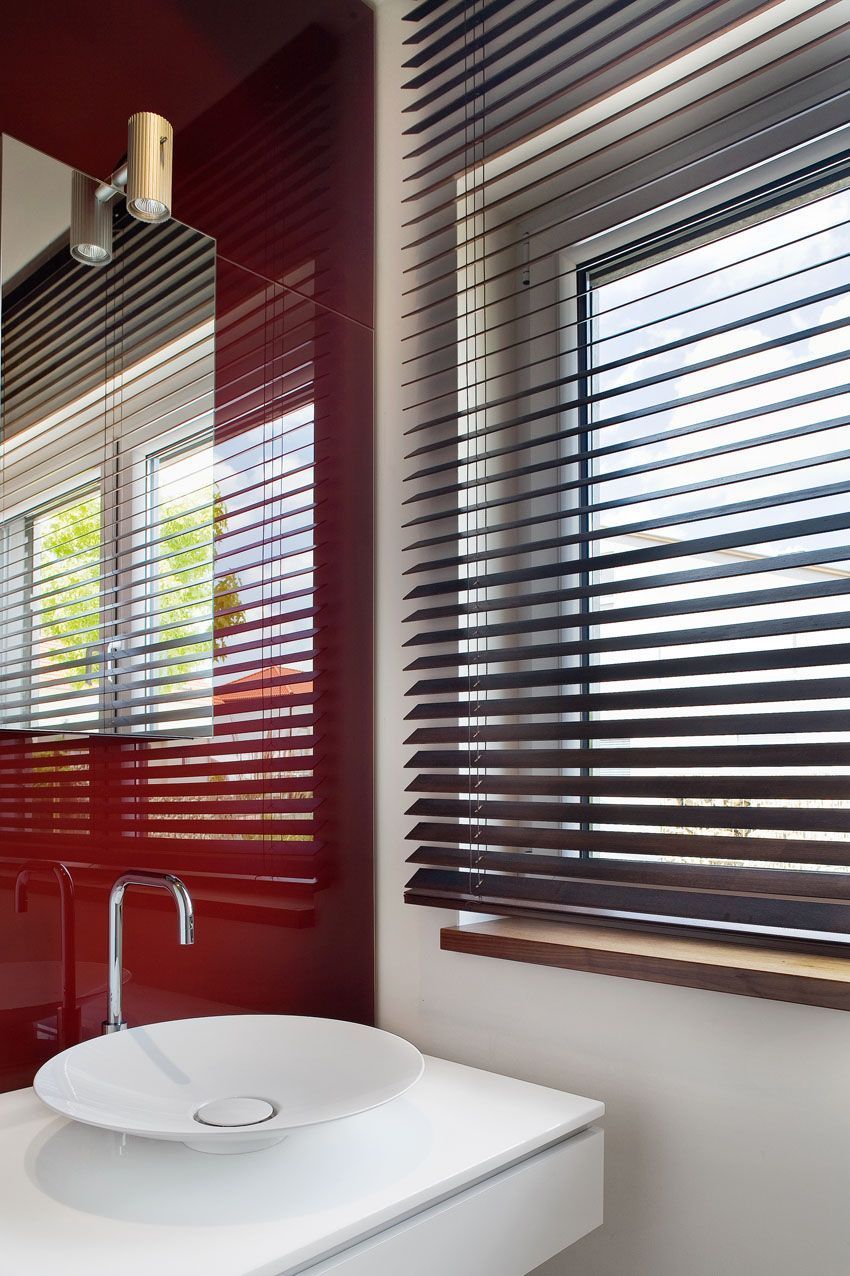 Badezimmer mit roten Wänden, weißem Waschbecken und dunklen Holzjalousien am Fenster.