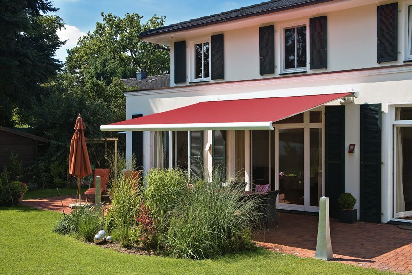 Eine rote Markise spendet Schatten über der Terrasse eines weißen Hauses mit schwarzen Fensterläden, das von grünem Gras und Pflanzen umgeben ist.
