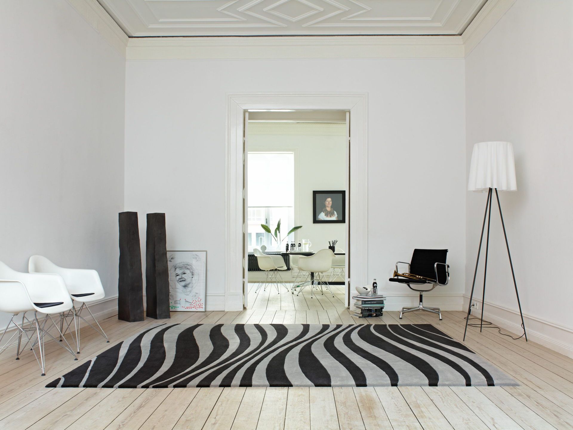 Weißes Zimmer mit gewelltem, schwarz-grauem Teppich, weißen Stühlen und schwarzer Stehlampe. Durch eine Tür gelangt man in einen anderen Raum.