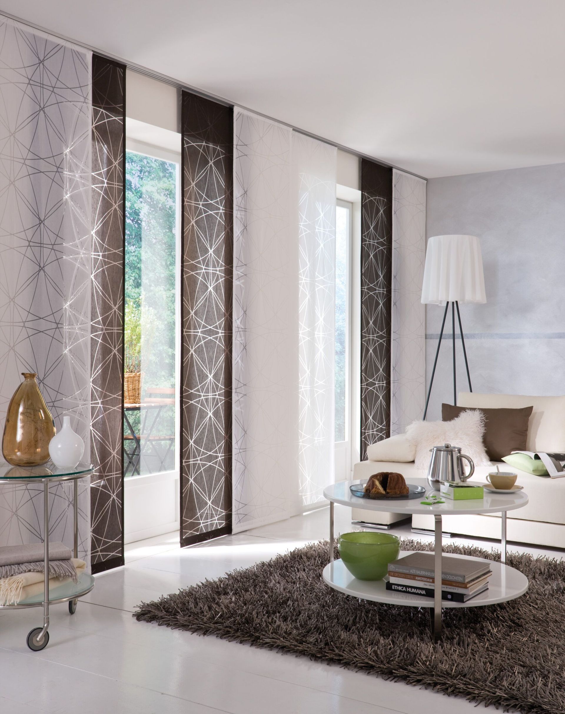 Wohnzimmer mit transparenten Flächenvorhängen, einem weißen Sofa und einem runden Couchtisch auf einem braunen Teppich.