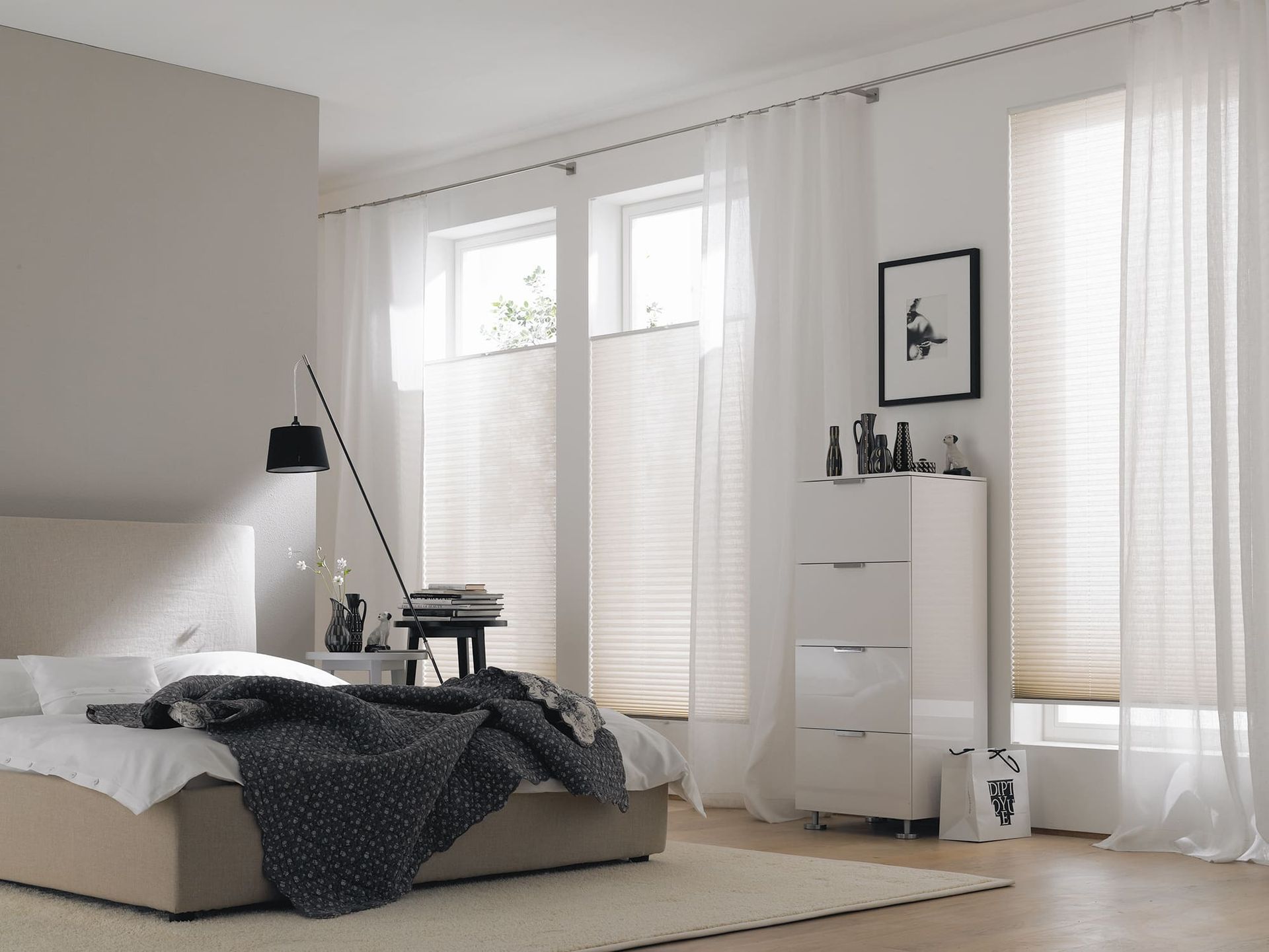 Schlafzimmer mit Bett, weißen Vorhängen und Plissees; weiße Kommode.