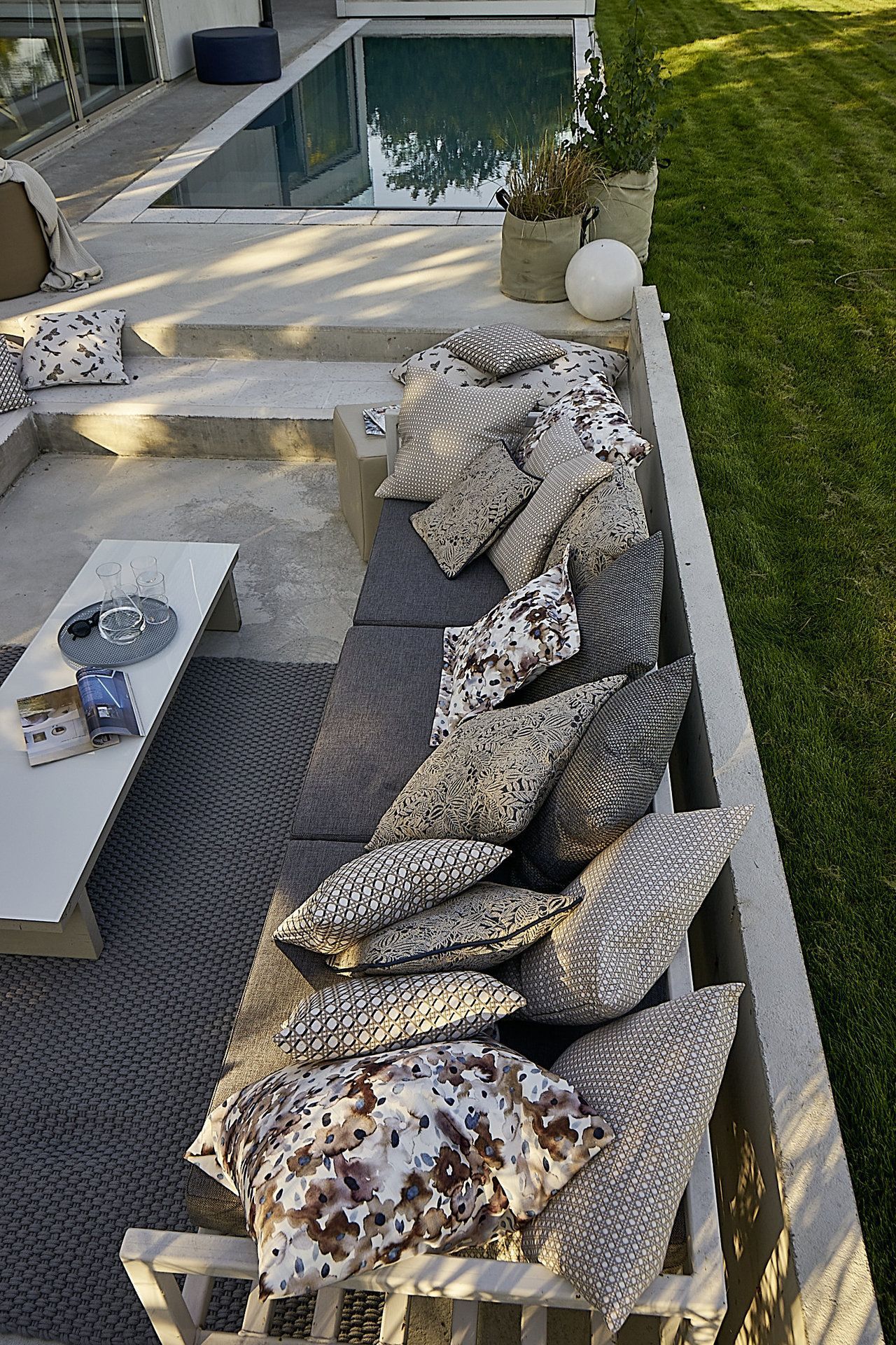 Sitzbereich im Freien mit grauen Kissen und Polstern, neben einem Pool und einer Rasenfläche.