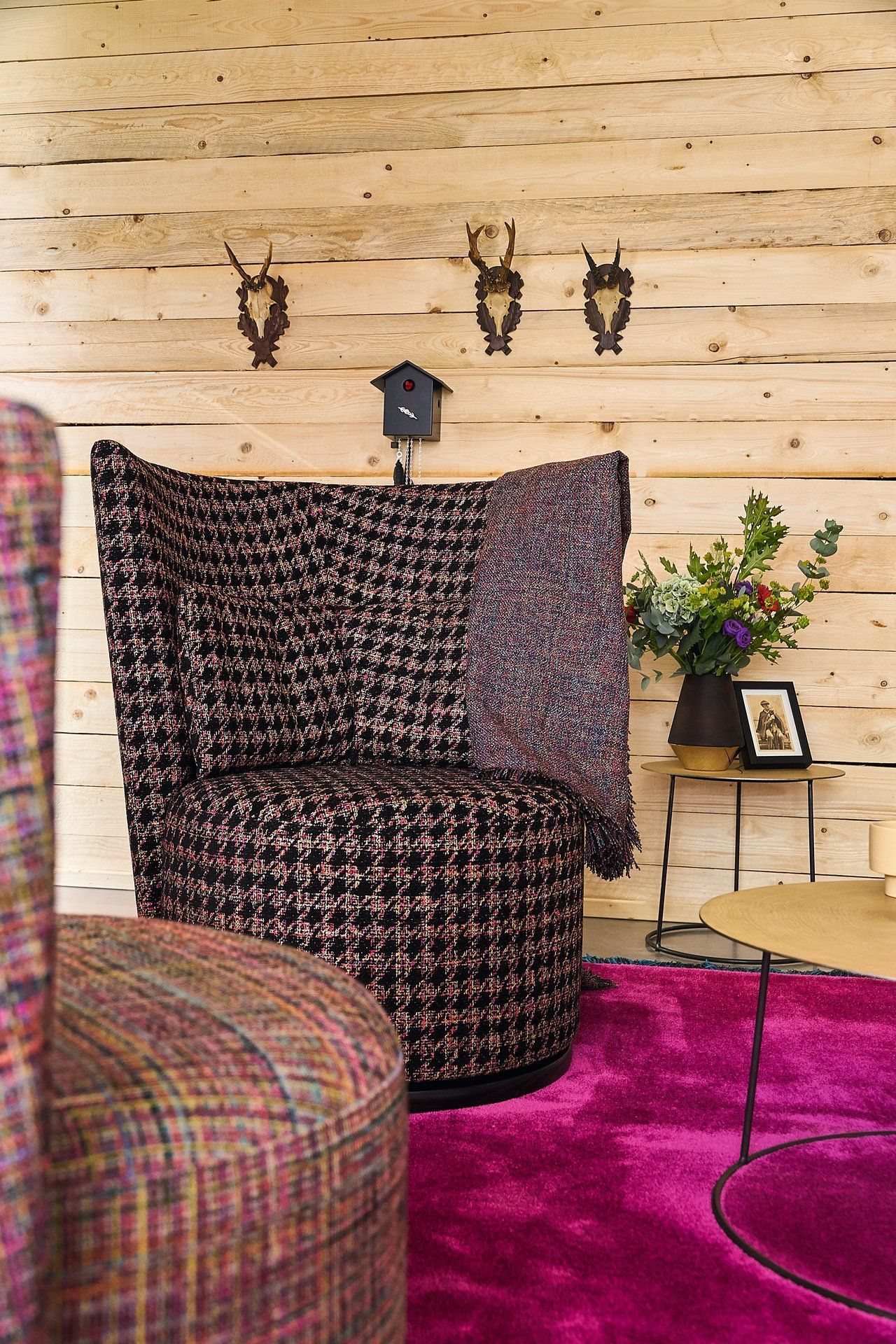 Gemütlicher Sessel mit gemustertem Stoffbezug, magentafarbener Teppich, Holzwand mit Hirschgeweihen, kleiner Tisch mit Blumen und Bilderrahmen.