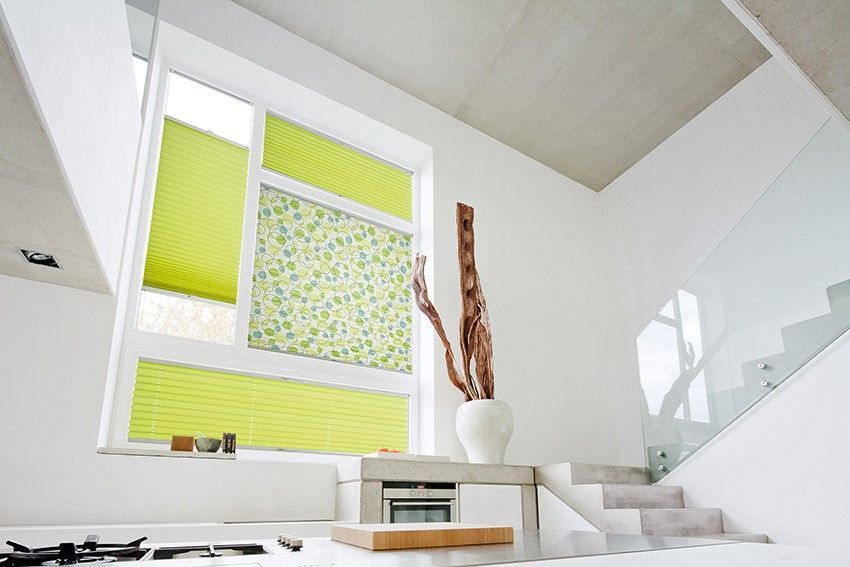 Modernes Interieur mit einem Fenster, das teilweise von grün gemusterten Jalousien verdeckt wird, einer weißen Vase mit Zweigen und einer Betontreppe.